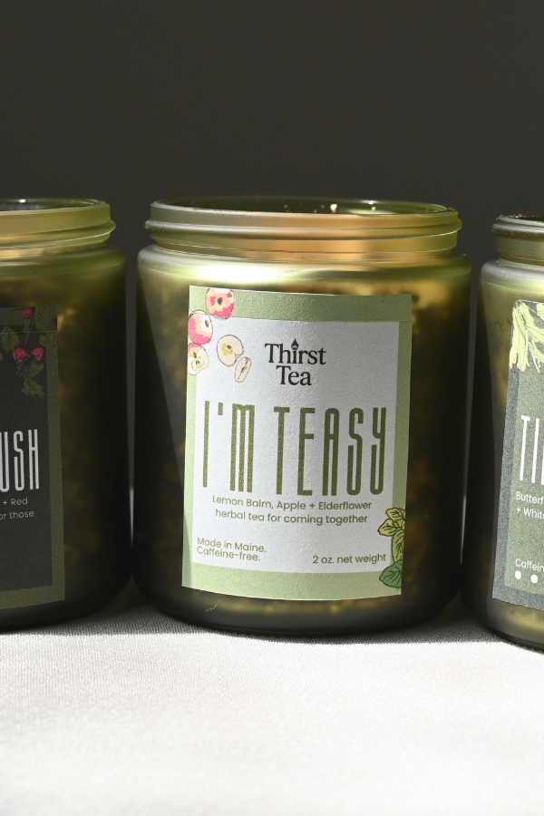 Three glass jars of Thirst Tea candles in a row on a light gray surface against a dark gray background. The label reads , 'I'M TEASY'