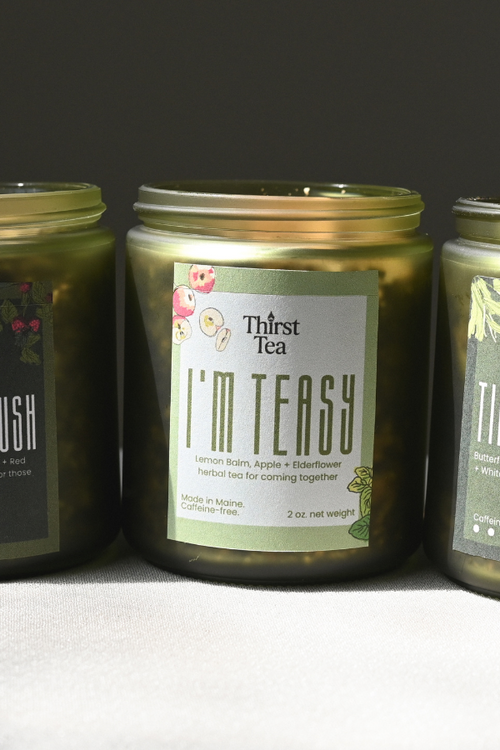 Three glass jars of Thirst Tea candles in a row on a light gray surface against a dark gray background. The label reads , 'I'M TEASY'